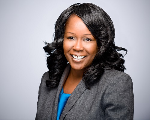 Headshot on a silver background of a smiling professional black woman wearing a blue blouse and grey business jacket, and long spiraling hair